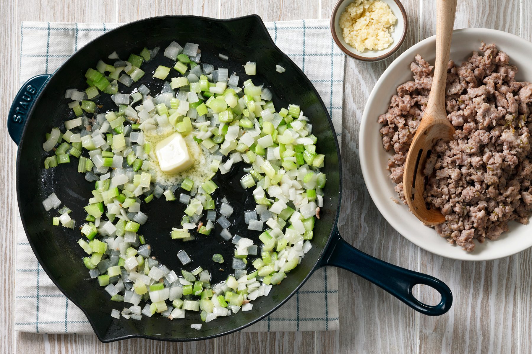Overhead shot of a cast iron skillet with diced onions and celery being cooked with a pat of butter placed over a white checkered dishtowel, There is a wooden spoon in a bowl of ground meat in the background.