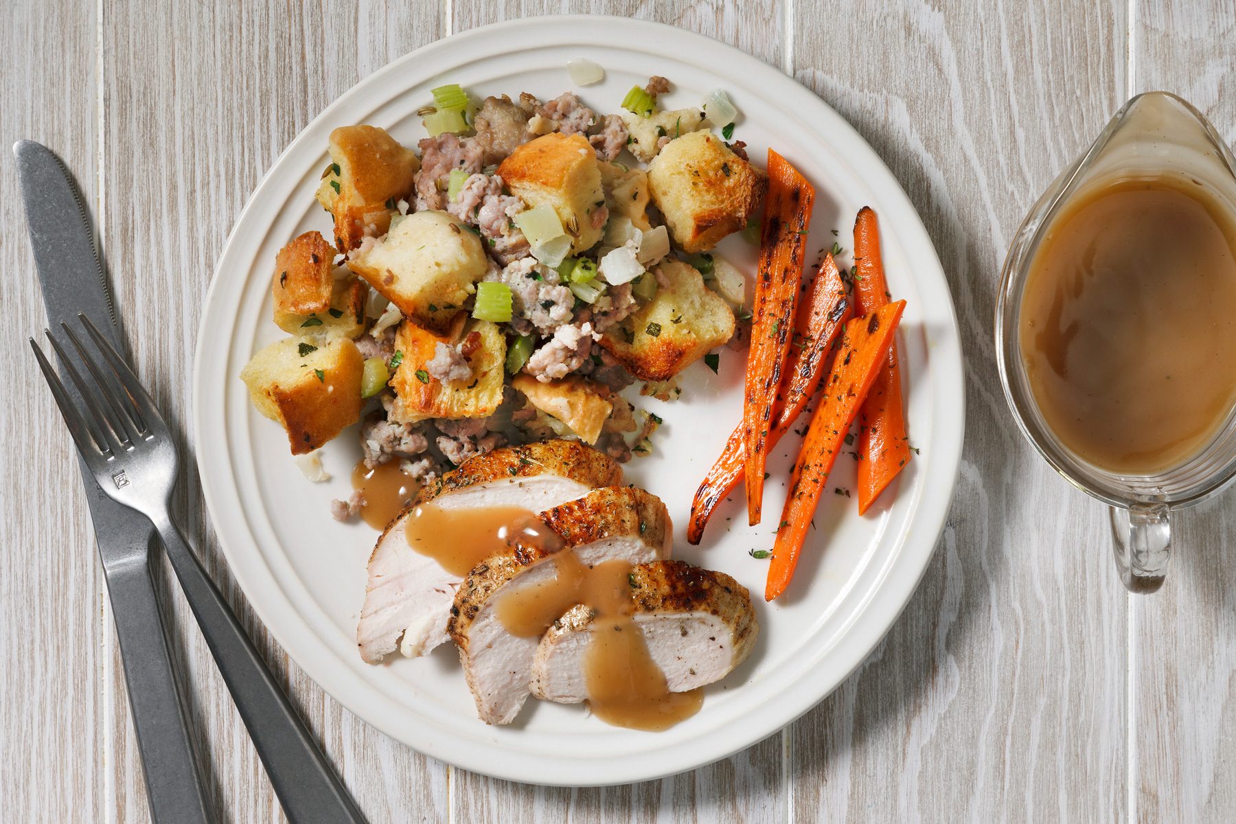 Overhead shot of a plate of food on a white wooden table; the plate has a white background and a white rim and it contains a variety of foods including Sausage Stuffing and carrots; there is also a gravy boat to the right of the plate which is filled with gravy; a knife and fork placed beside the table.