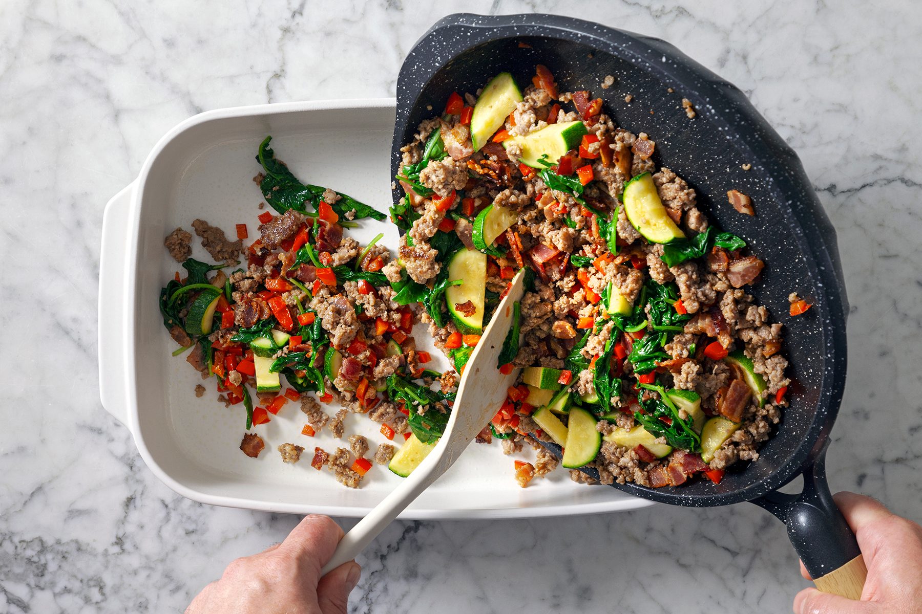 Hands transferring cooked ground meat, zucchini, spinach, and bell peppers from a black skillet into a white baking dish on a marble countertop.