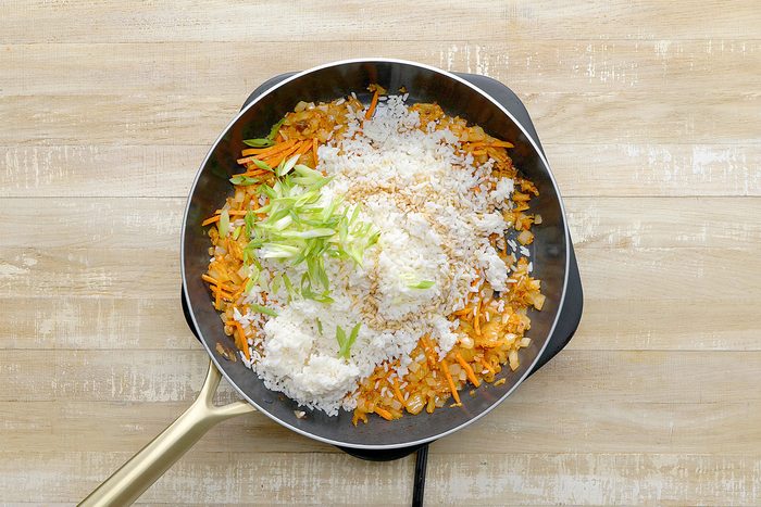 overhead shot of a black skillet contains a mixture of sautéed vegetables, kimchi, and cooked rice, The vegetables are a vibrant orange and white color, and the rice is a white color, Chopped green onions are scattered on top of the mixture;
