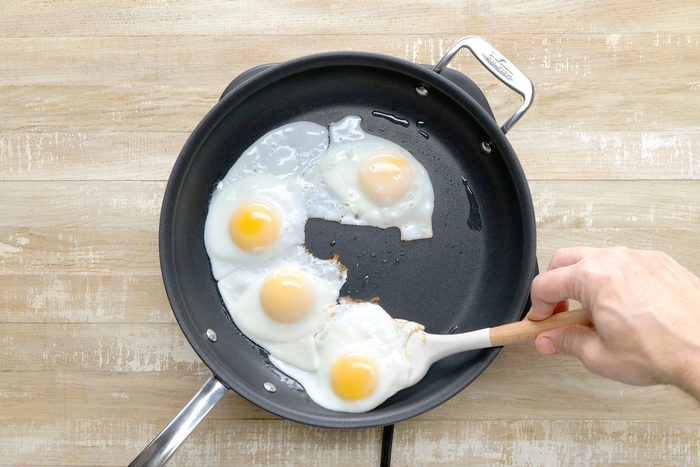 overhead shot of a non-stick skillet filled with oil, Four fried eggs are cooking in the oil, with the yolks still runny, A wooden spatula is being used to gently lift one of the eggs, revealing the cooked white underneath;