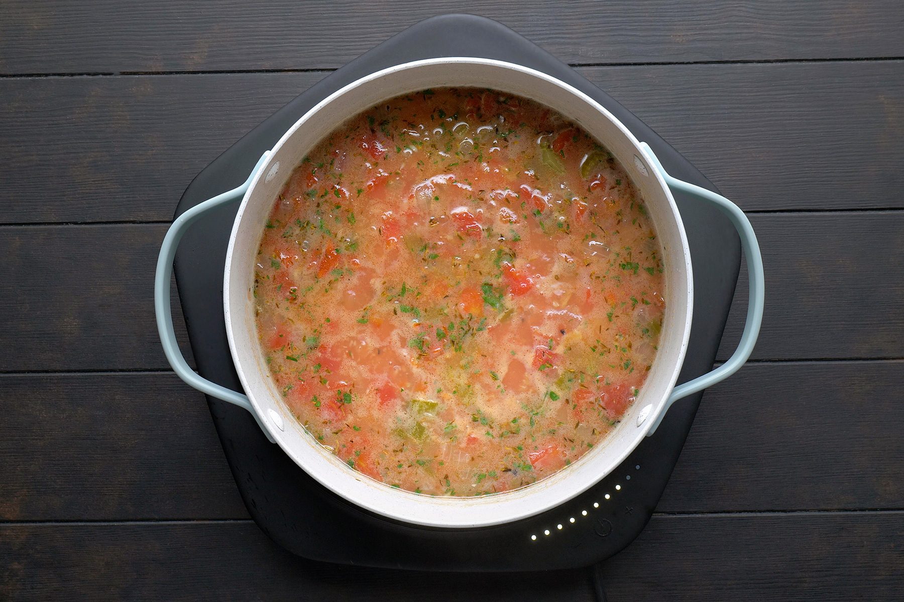 A pot of soup on a black cooking surface, containing a mix of chopped vegetables such as tomatoes, herbs, and other ingredients, creating a colorful and hearty dish. The pot has light-colored handles.