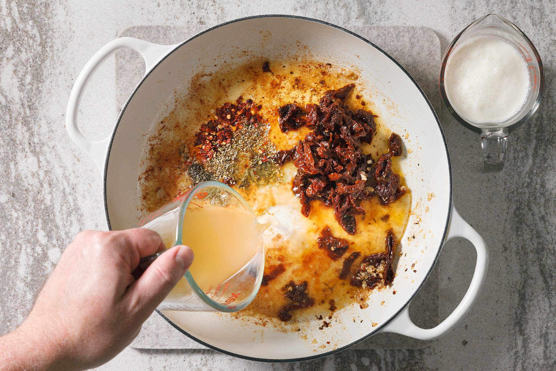 overhead shot of a white pot with a black rim. The pot is filled with a mixture of oil, herbs, spices, and sun-dried tomatoes, A hand is holding a glass measuring cup filled with a yellowish liquid and pouring it into the pot, There is another glass measuring cup in the upper right corner, filled with a white liquid, The background is a speckled gray countertop;