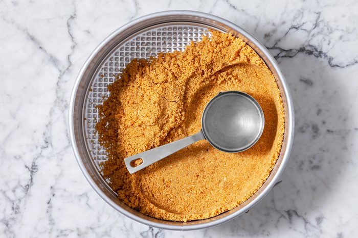 Overhead shot of a pie crust being prepared in a springform pan, The crust is made of crushed graham crackers, which are a light brown color, The crust is pressed firmly into the bottom of the pan and is ready to be filled with a creamy filling.