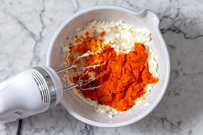 Overhead shot of a white bowl containing a mixture of cream cheese and pumpkin puree, An electric mixer is partially submerged in the mixture.