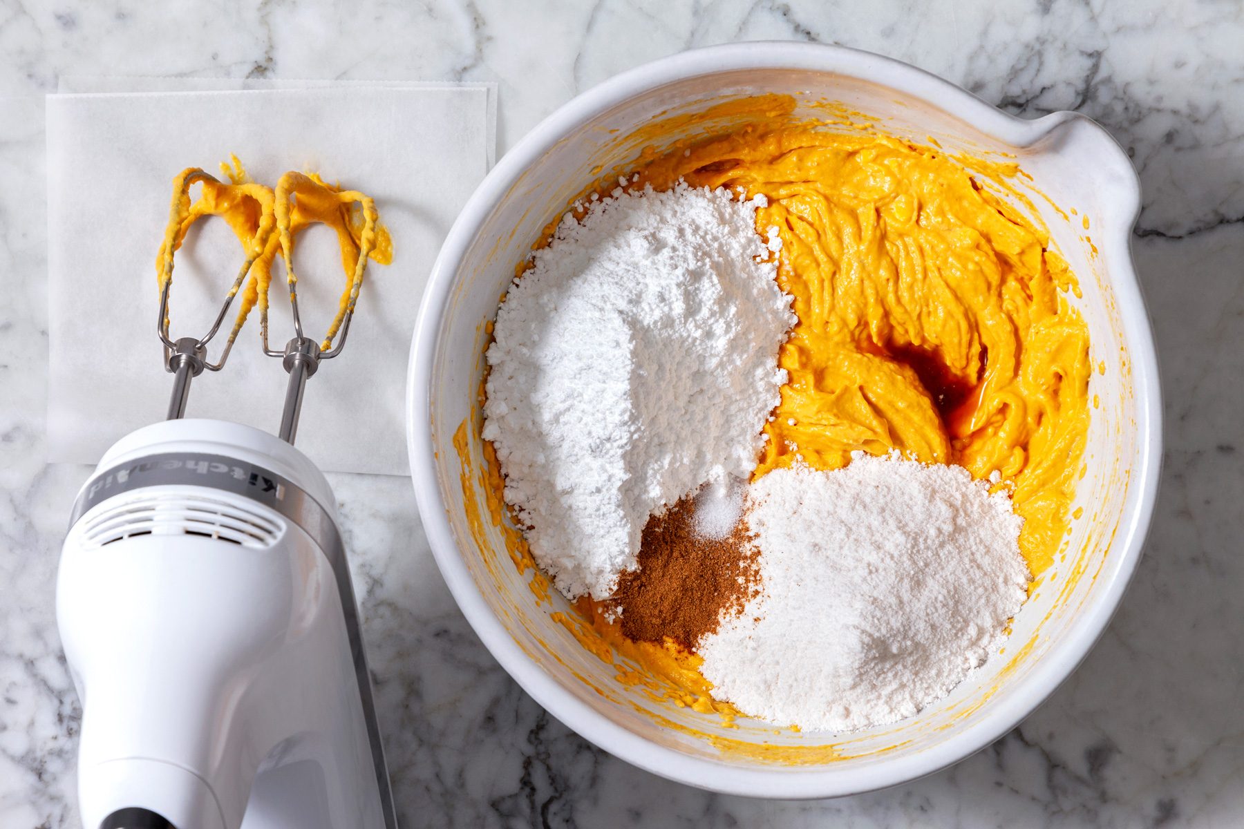 Overhead shot of a white bowl containing a mixture of pumpkin puree, cream cheese, and powdered sugar next to an electric mixer partially submerged in the pumpkin mixture rests on a napkin.