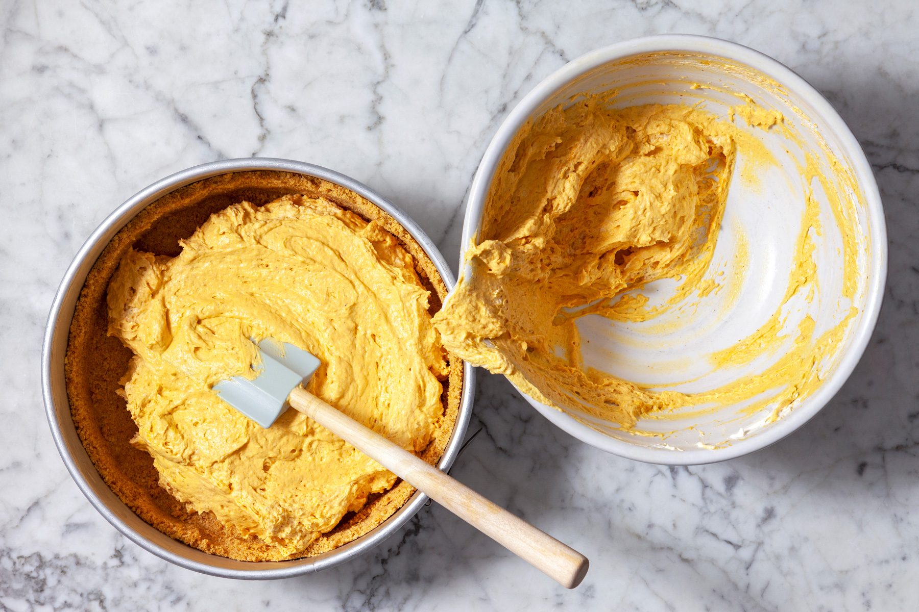 Overhead shot of a springform pan filled with a pumpkin cheesecake batter, The batter is a vibrant orange color and is being spread evenly with a spatula, A second bowl contains the remaining batter, which is being scooped out with a wooden spoon.