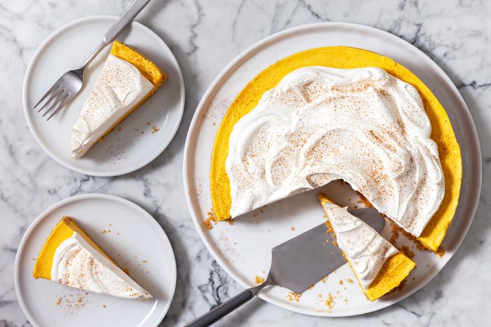 Overhead shot of a pumpkin cheesecake on a white plate, The cheesecake is a vibrant orange color and is topped with a layer of whipped cream, A sprinkle of cinnamon is dusted over the whipped cream, Three slices of cheesecake are cut from the whole cheesecake and are placed on smaller plates, A fork and a server are placed next to the cheesecake.