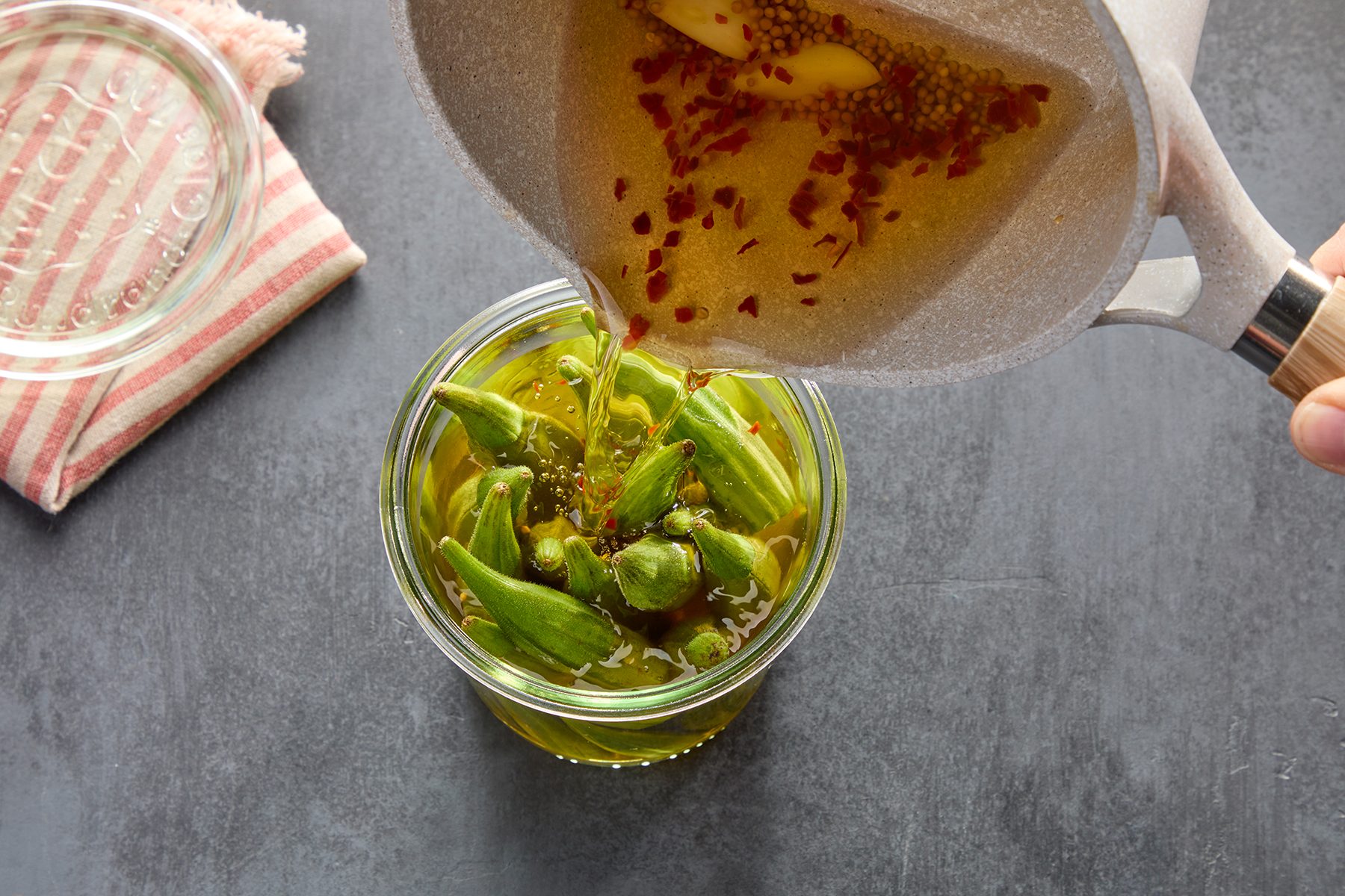 A hand pours a spiced brine from a pot into a jar filled with fresh okra. A red-striped cloth and a lid are nearby on a dark surface. The brine contains visible crushed red pepper and mustard seeds.