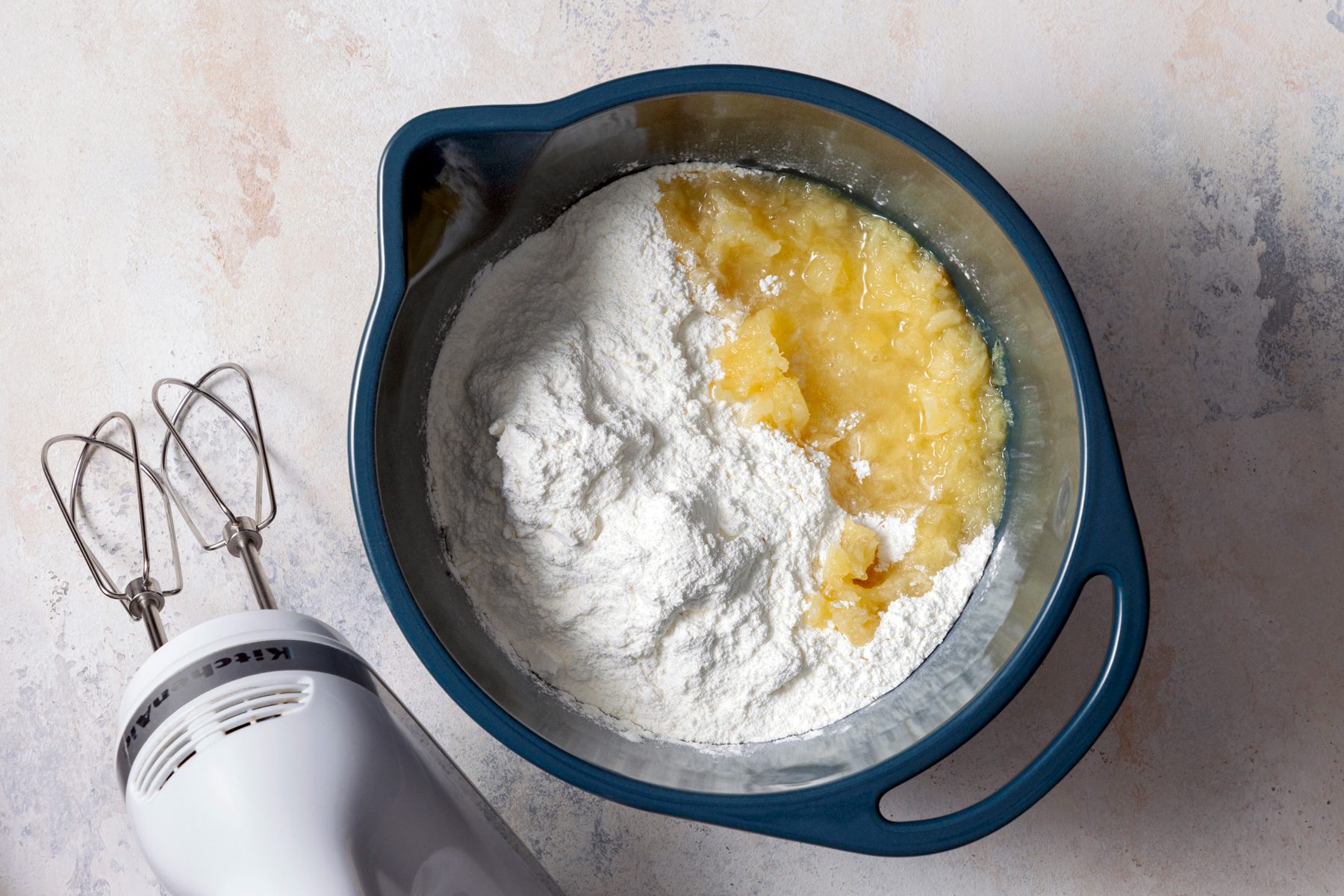 A large mixing bowl contains a mixture of angel food cake mix and crushed pineapple with juice. The ingredients are being beaten together on medium-low speed until fully combined.