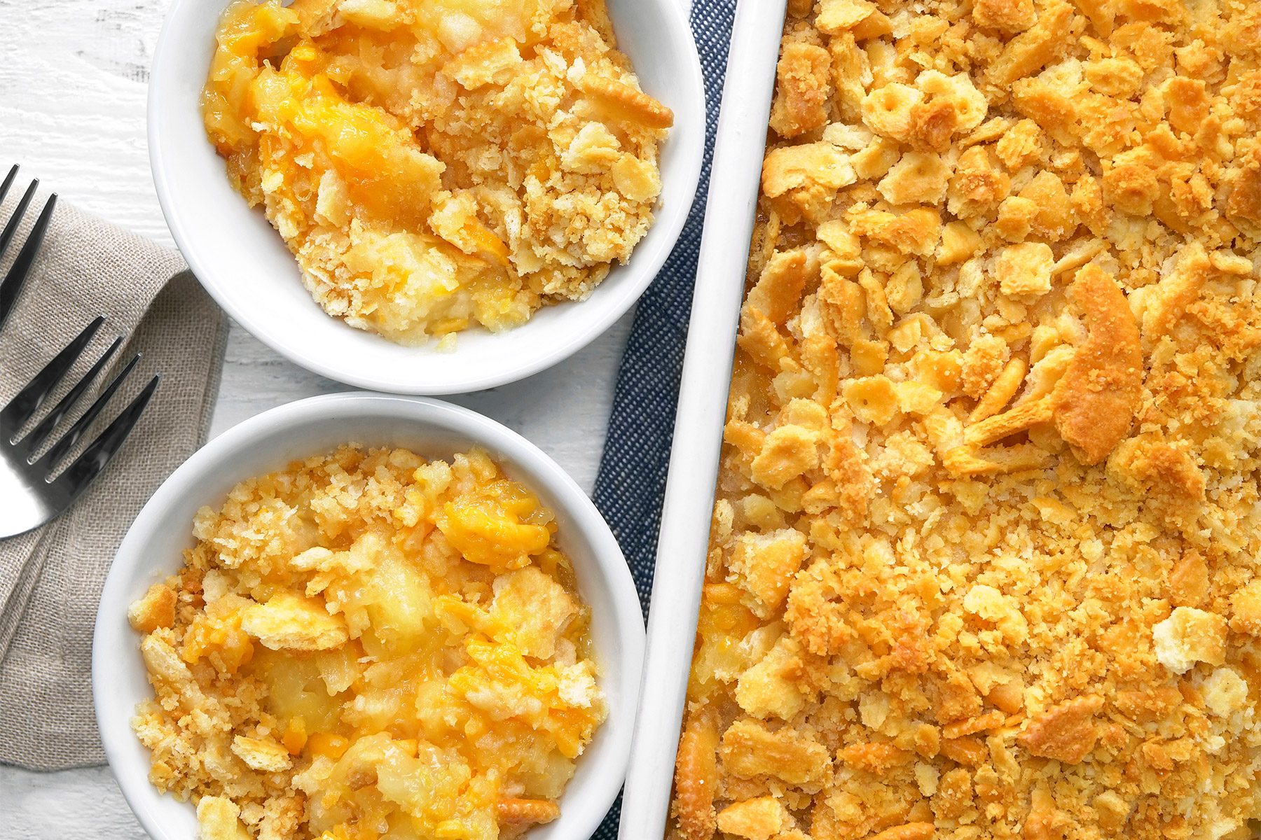overhead shot of a Pineapple Casserole, It features a large, rectangular baking dish and a smaller, round serving bowl, both containing the casserole, The casserole has a golden-brown crumbly topping with visible chunks of pineapple underneath. two silver forks placed aside over kitchen cloth;