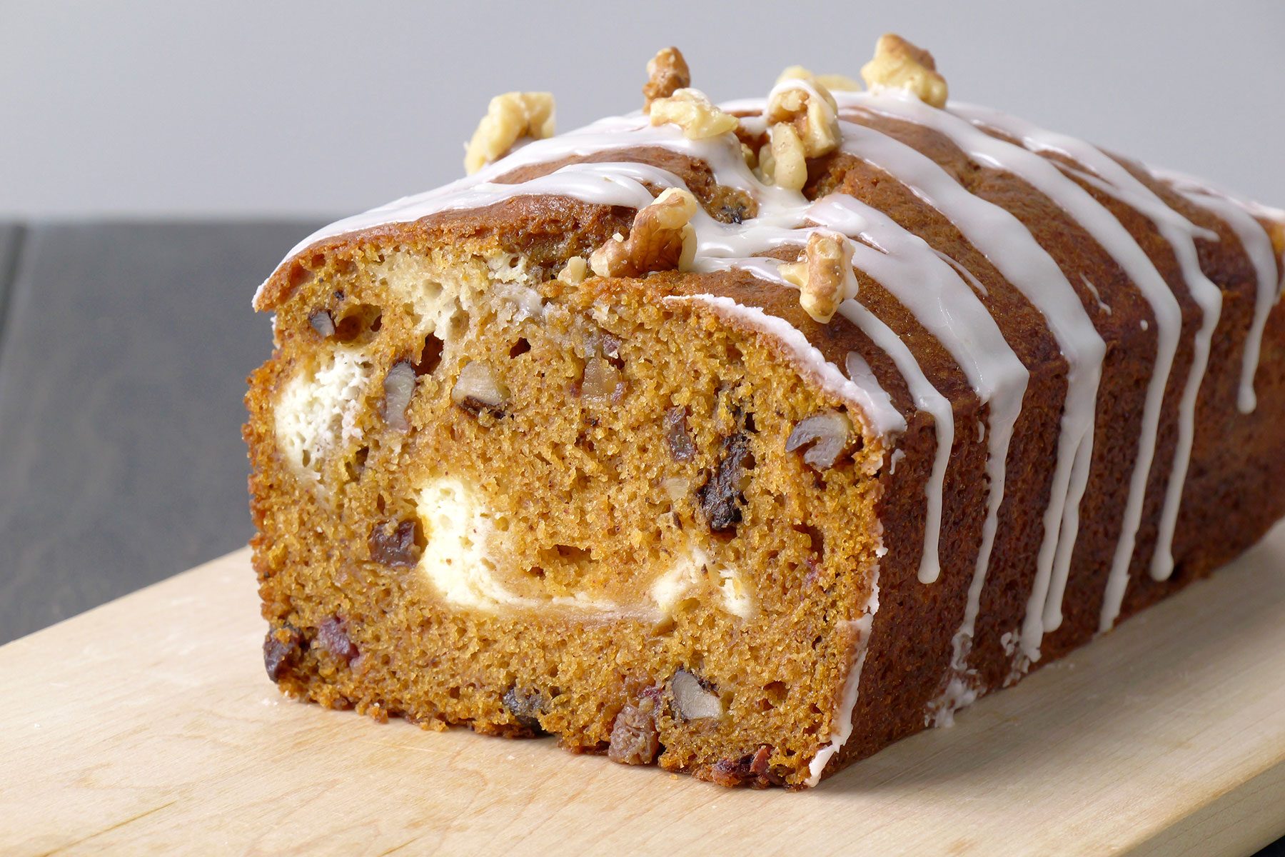 A loaf of pumpkin bread with cream cheese filling, sliced to reveal its interior. The top is drizzled with white icing and sprinkled with chopped walnuts, resting on a wooden cutting board.