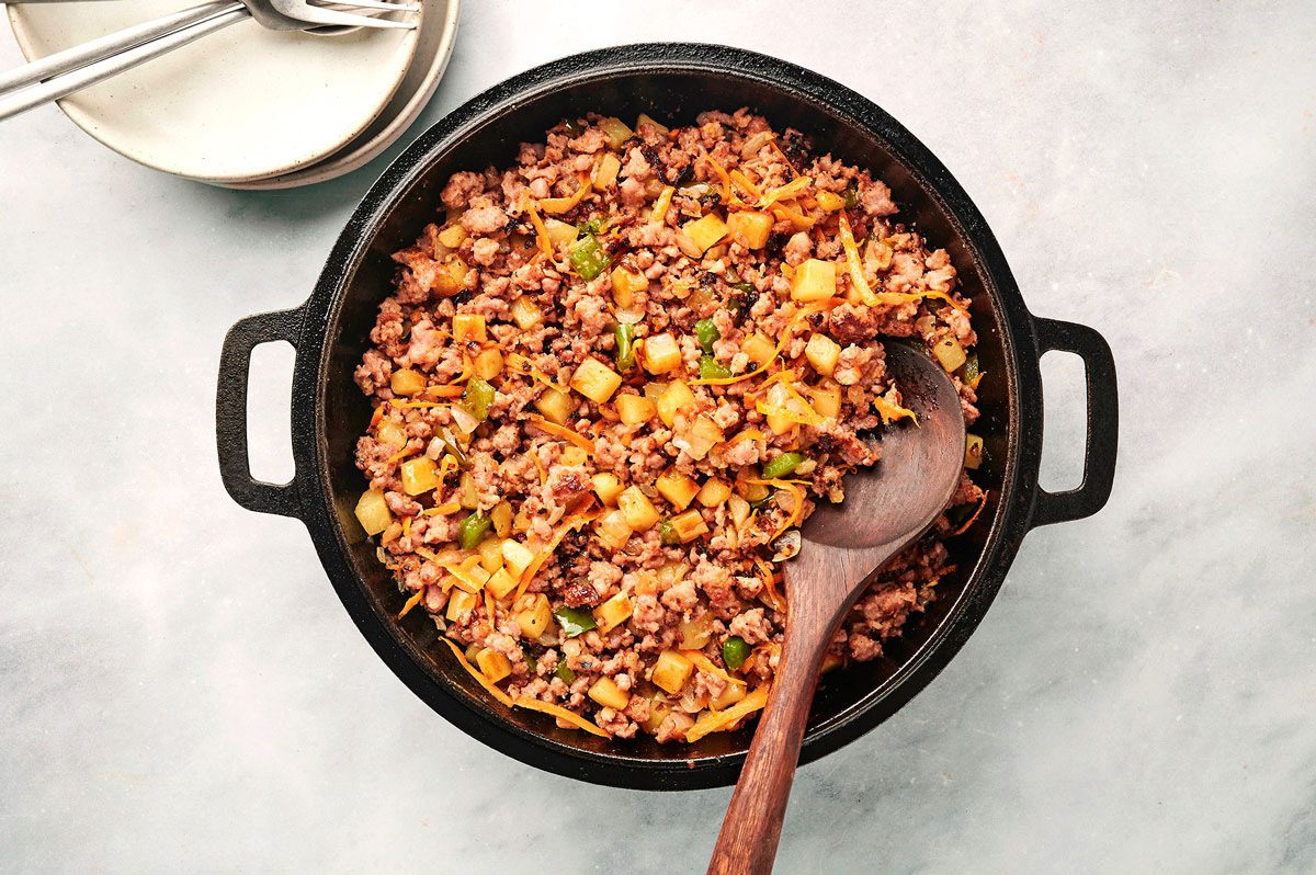 Adding veggies to browned sausage in a cast iron skillet and cooking the Sausage Hash recipe by Taste of Home