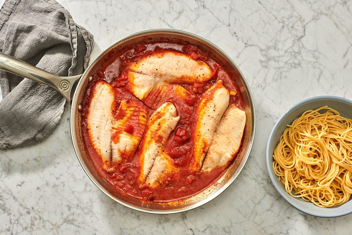 Cooked fish and sauce in a skillet with pasta nearby