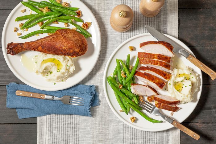 Two plates on a table: one with a roasted turkey leg, mashed potatoes, and green beans; the other with sliced turkey breast, mashed potatoes, and green beans. A fork and knife are by the plates, and two pepper mills are in the background.