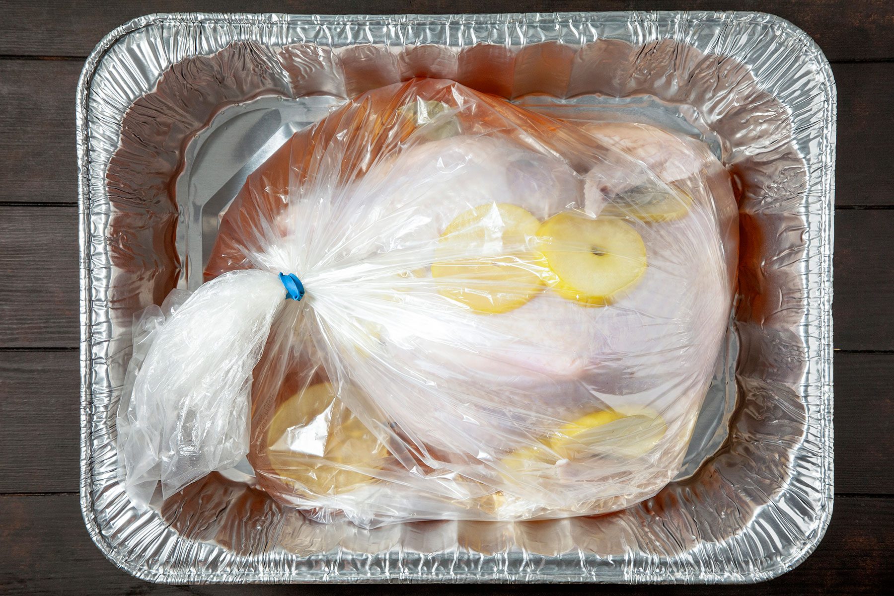A large raw turkey is placed in a plastic cooking bag with lemon slices. The bag is sealed with a blue tie and rests in a rectangular aluminum roasting pan on a dark wooden surface.