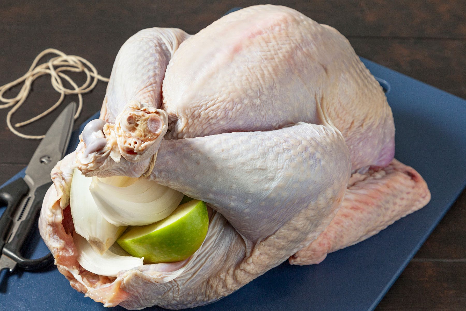 A raw whole turkey on a dark cutting board with its cavity filled with onion and apple pieces. Kitchen scissors and twine are nearby, suggesting preparation for roasting.