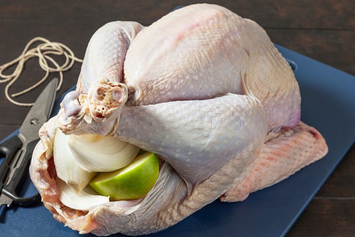 A raw whole turkey on a dark cutting board with its cavity filled with onion and apple pieces. Kitchen scissors and twine are nearby, suggesting preparation for roasting.