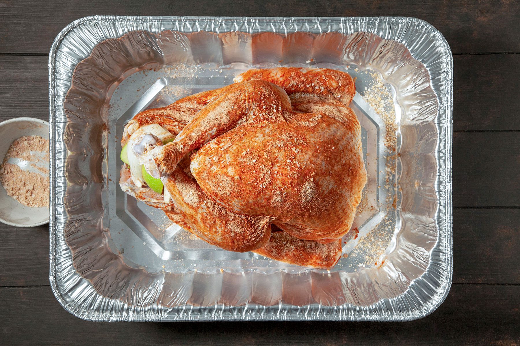 A raw, seasoned whole turkey placed in a large, rectangular aluminum roasting pan, ready for cooking. The turkey is sprinkled with spices, and a small bowl of seasoning is visible beside the pan on a dark wooden surface.