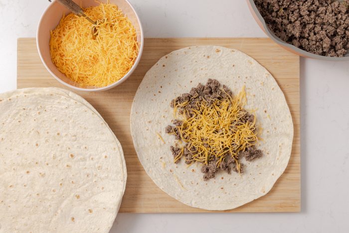 Burritos being assembled with ground beef mixture and cheddar cheese.