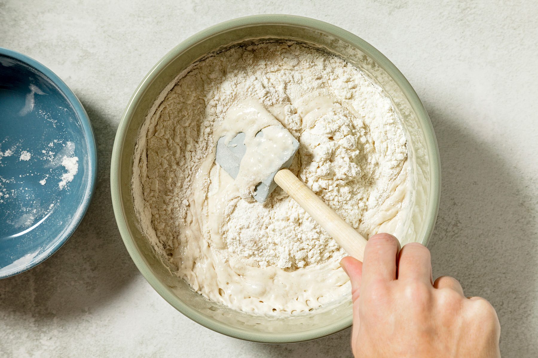 overhead shot of a green bowl contains a mixture of flour and a wet, bubbly substance, A hand is using a spatula to gently fold the flour into the mixture, The mixture appears to be a light yellow color and has a slightly sticky consistency;
