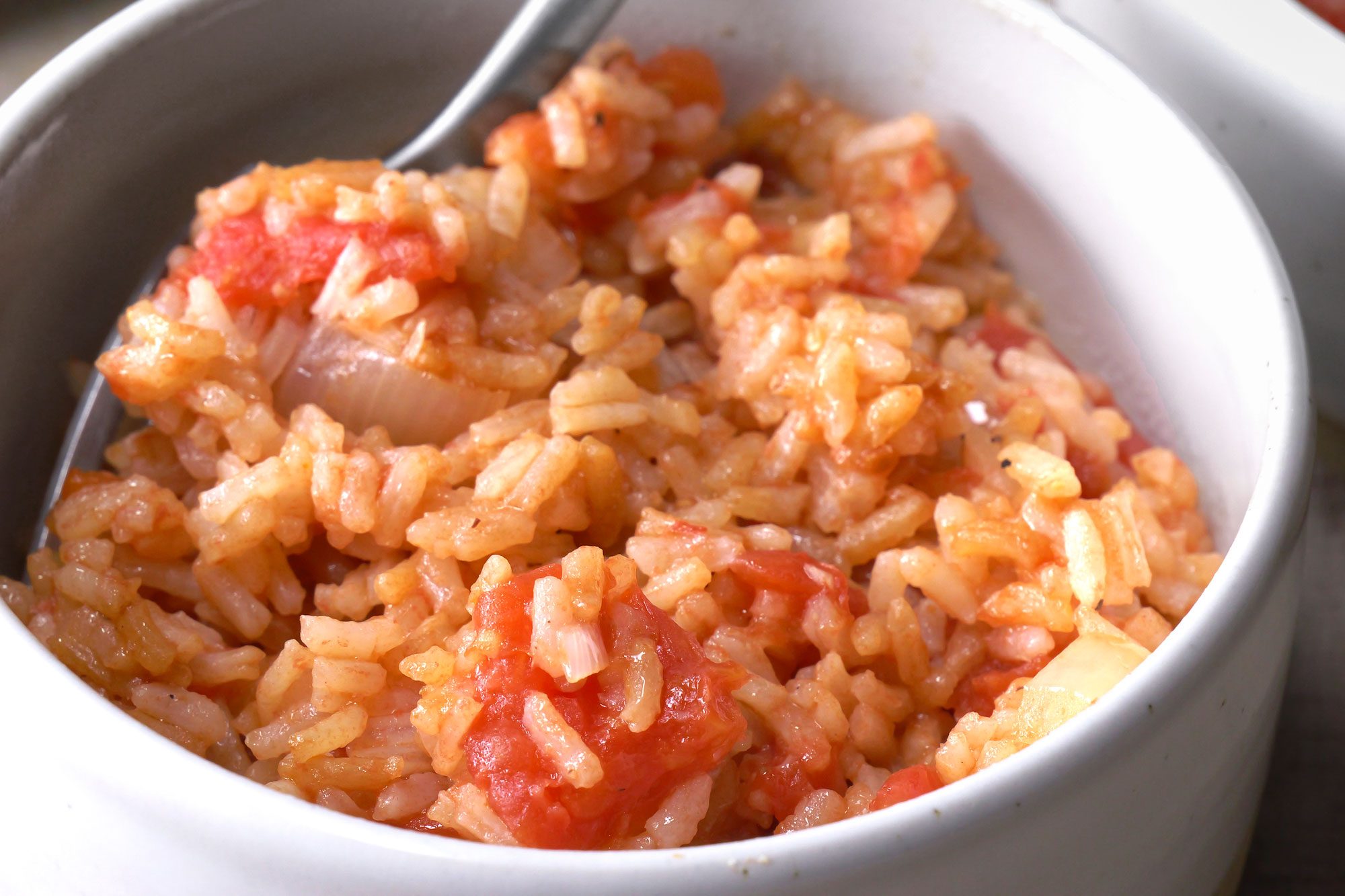 Bowl of rice that is red in color and has a slightly grainy texture, The rice is mixed with chopped tomatoes, onions, and other vegetables. A spoon is resting in the bowl;