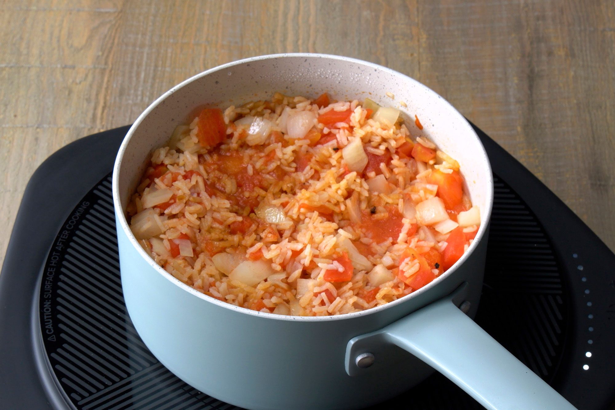 The saucepan contains a mixture of rice, chopped tomatoes, and chopped onions, The rice is a white color and the tomatoes and onions are a vibrant red color, The mixture is slightly oily