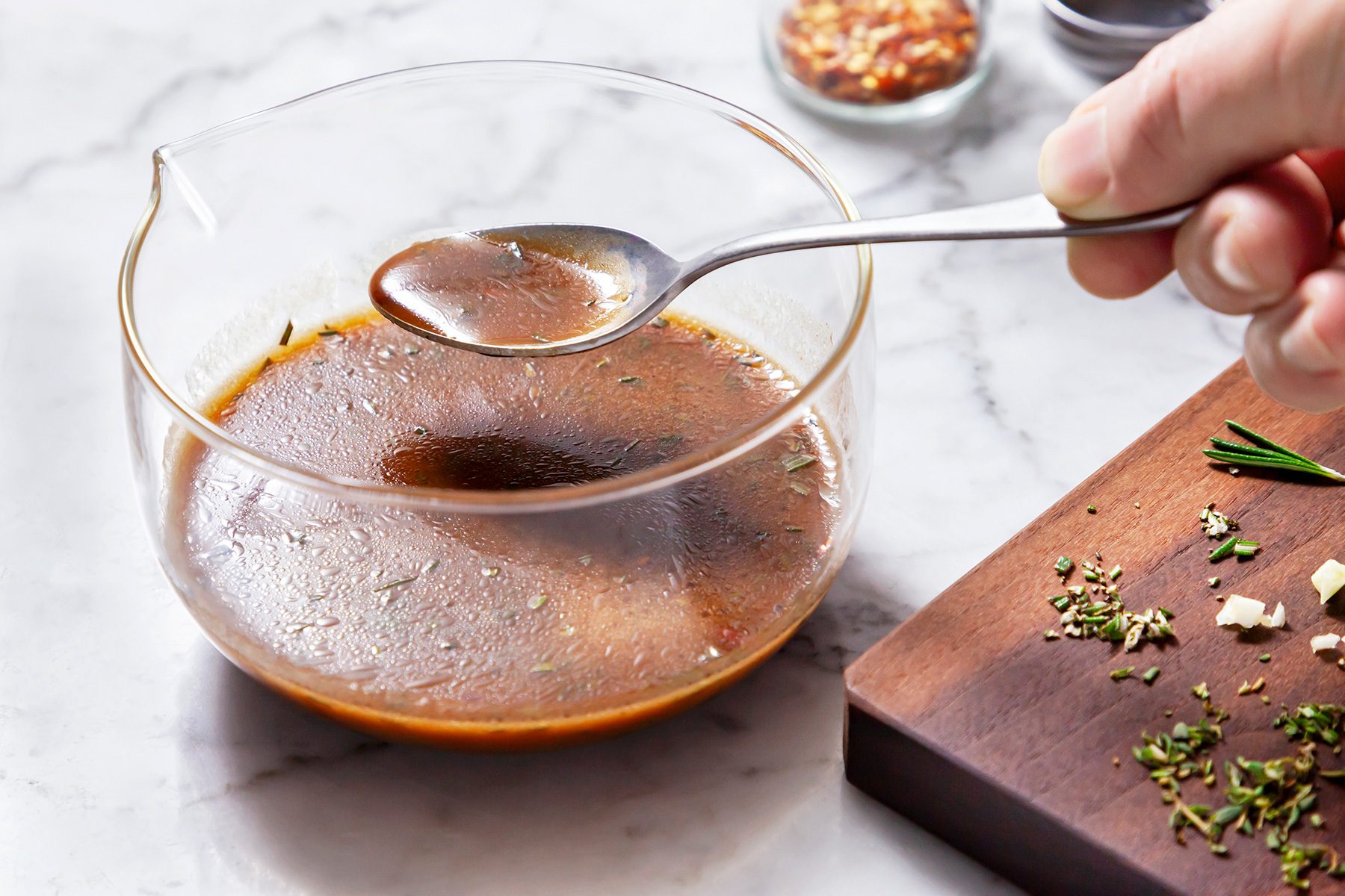 3/4th vertical shot of a glass bowl filled with a rich, dark marinade or sauce, set against a marble background, A long-handled metal spoon rests in the mixture;