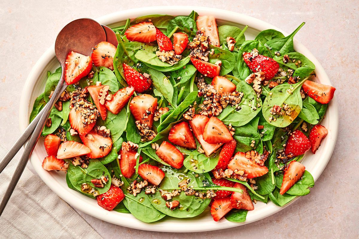 A white serving platter with the strawberry spinach salad recipe by Taste of Home, with two metal serving spoons