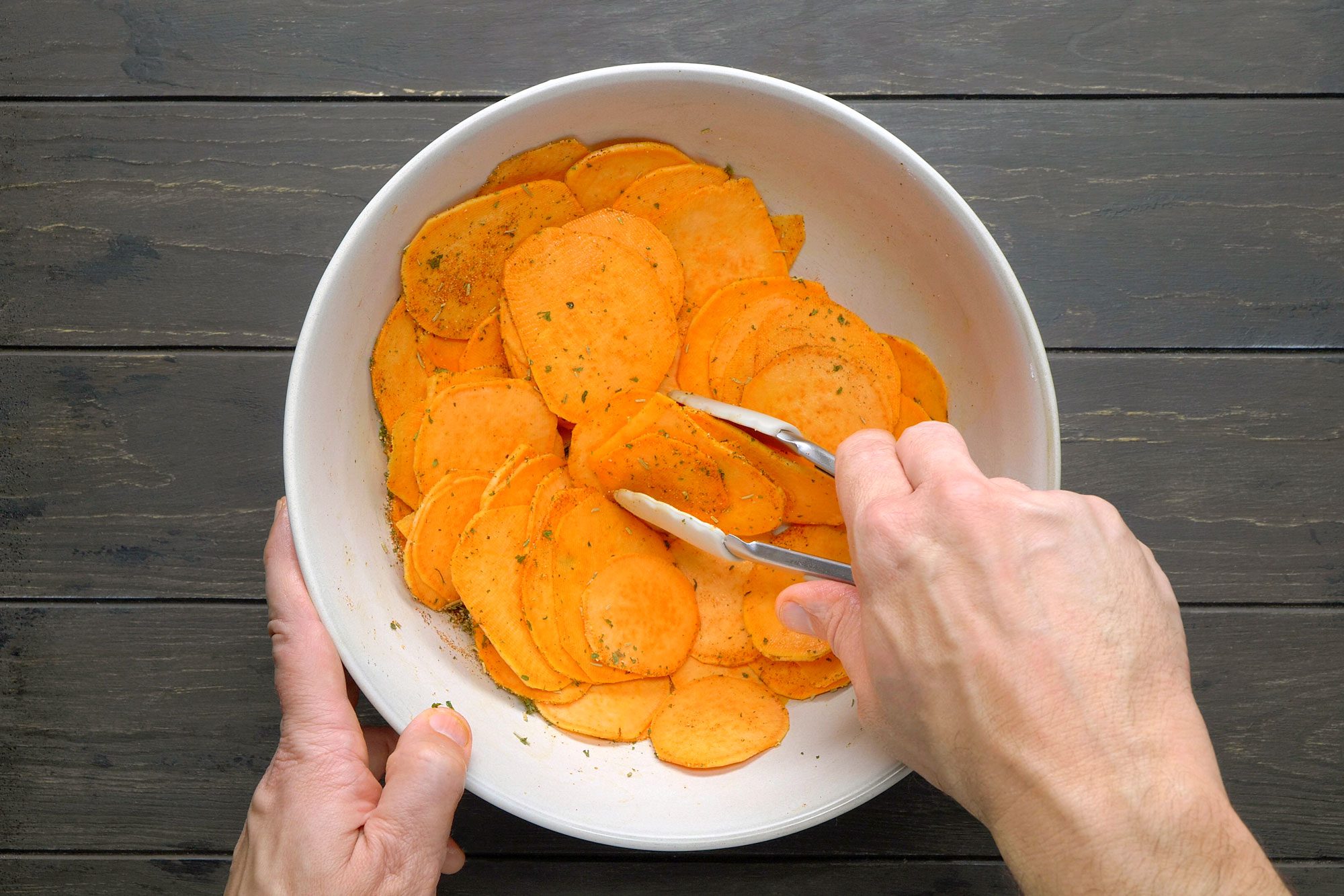 White bowl filled with sliced sweet potatoes and tossing with tongs