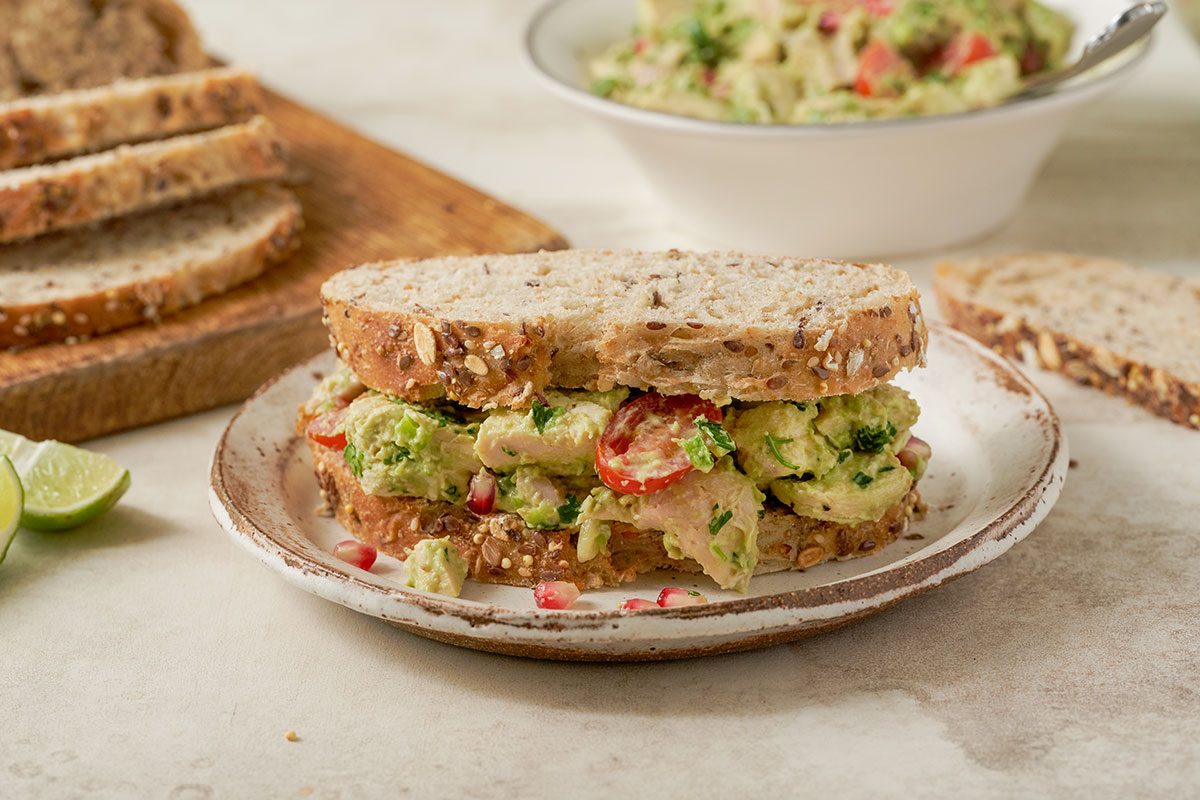 Close-up horizontal shot of the Taste of Home Avocado Chicken Salad sandwich, highlighting the creamy avocado, juicy cherry tomatoes, and tender chicken.