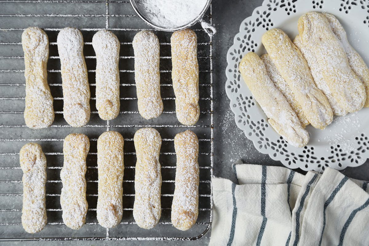 Close up of Taste of Home's Lady Fingers on a wire rack on a grey marble surface with powdered sugar.
