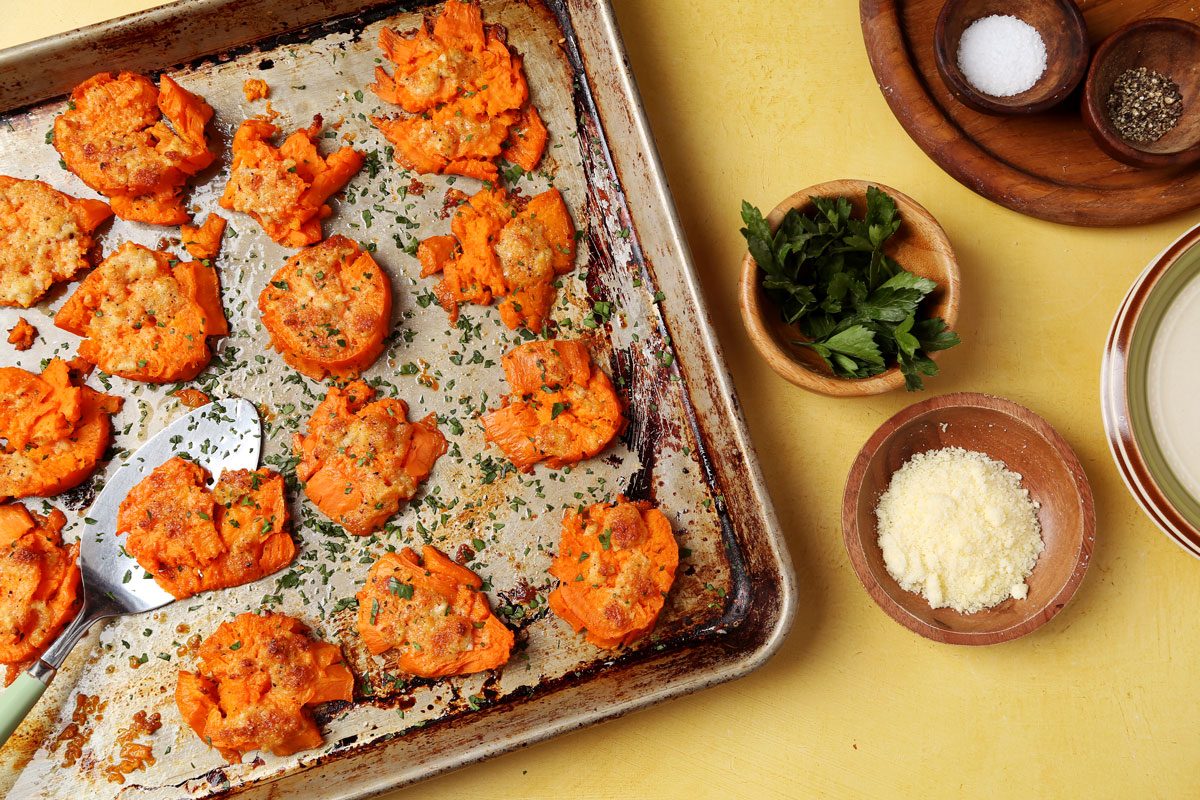 Close up of Taste of Home's Smashed Sweet Potatoes on a large baking sheet on a yellow surface.