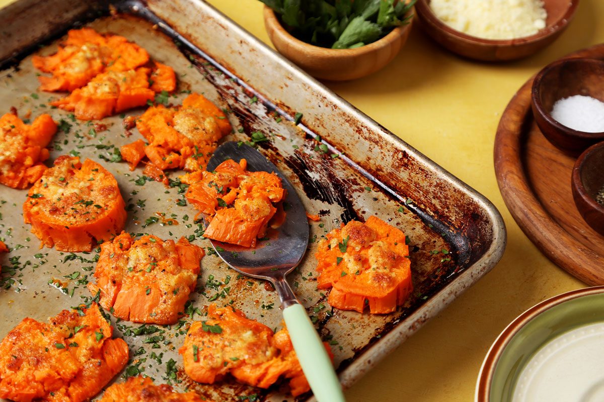 Close up of Taste of Home's Smashed Sweet Potatoes on a large baking sheet on a yellow surface.