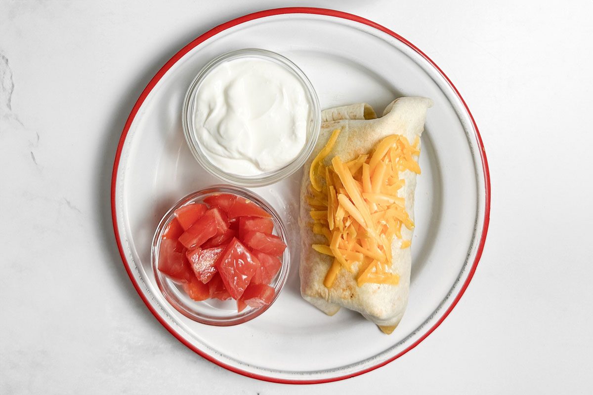 Taste of Home Taco Burrito on an enamel plate with bowls of sour cream and tomatoes