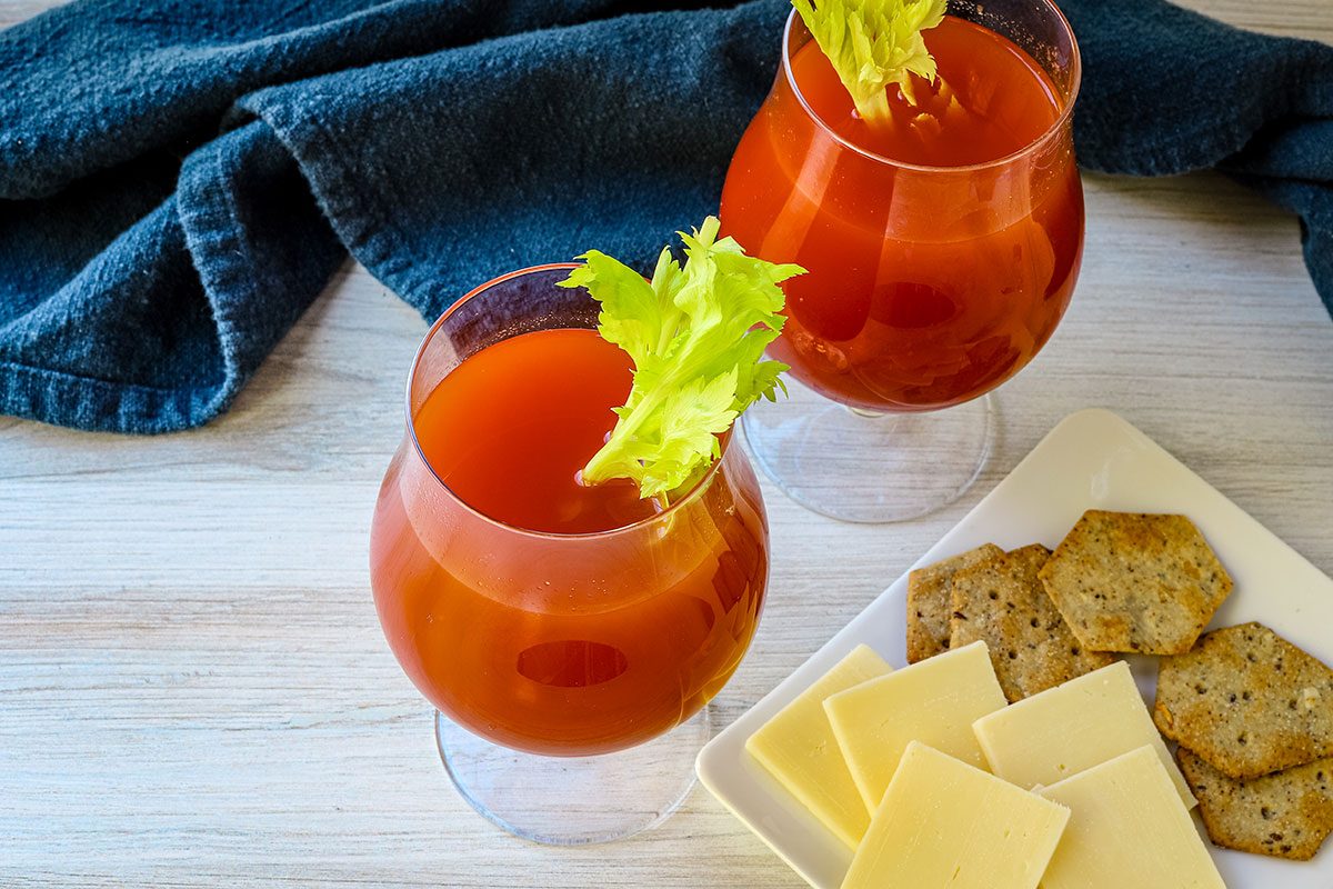 Homemade spicy tomato juice in glasses with celery and crackers and cheese alongside them.