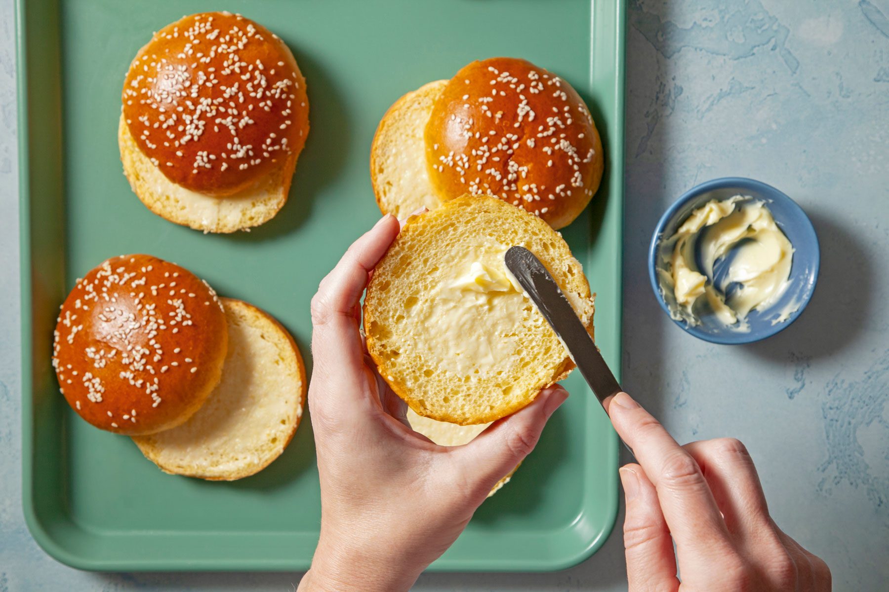 Hands can be seen spreading butter over the cut sides of buns.