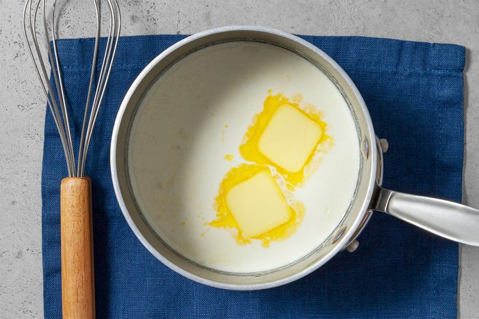 Overhead shot of a small saucepan heat cream and butter over medium heat until bubbles form around the edges; stirring occasionally; do not boil; blue cloth; whisk tool; grey surface;