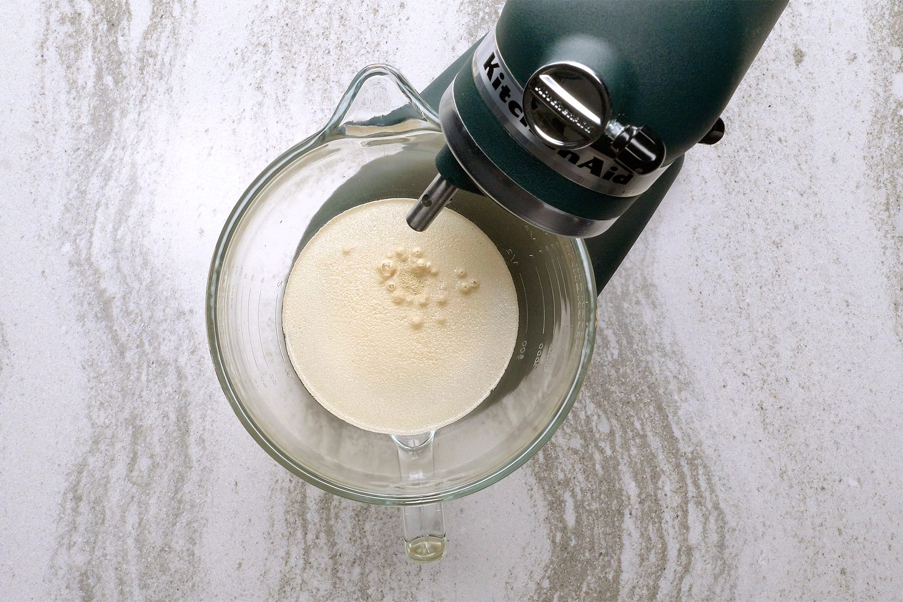 A top-down view of a stand mixer with a glass bowl attached. The bowl contains a light-colored creamy mixture, and the mixer is set on a marble countertop.