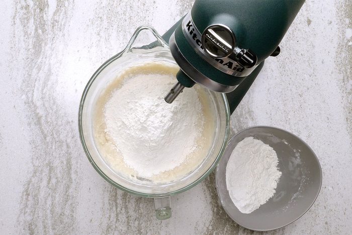 A green stand mixer with a glass bowl is mixing white ingredients on a marble countertop. A gray bowl with white powder is nearby.