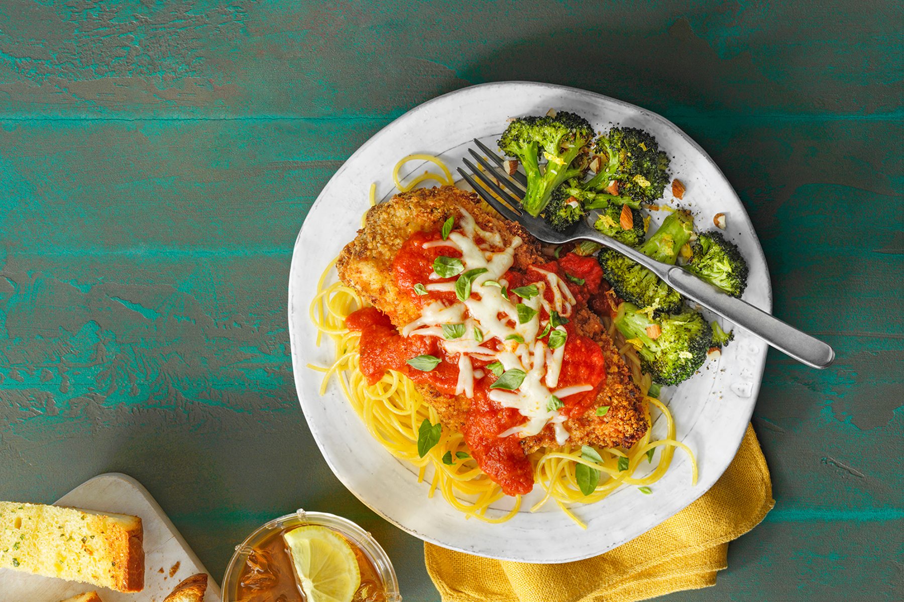 A plate of chicken parmesan topped with tomato sauce and melted cheese sits on a bed of spaghetti. Roasted broccoli is on the side, and a fork rests on the plate. There's a piece of bread and a glass of iced tea with lemon nearby on the table.