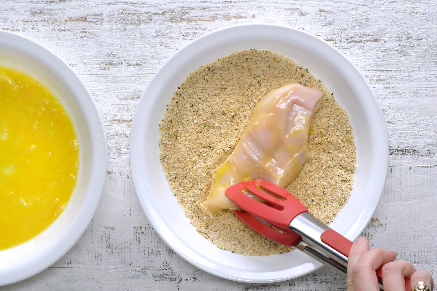 A person uses red tongs to dip a raw chicken breast in breadcrumbs, beside a bowl of beaten eggs on a white wooden surface.