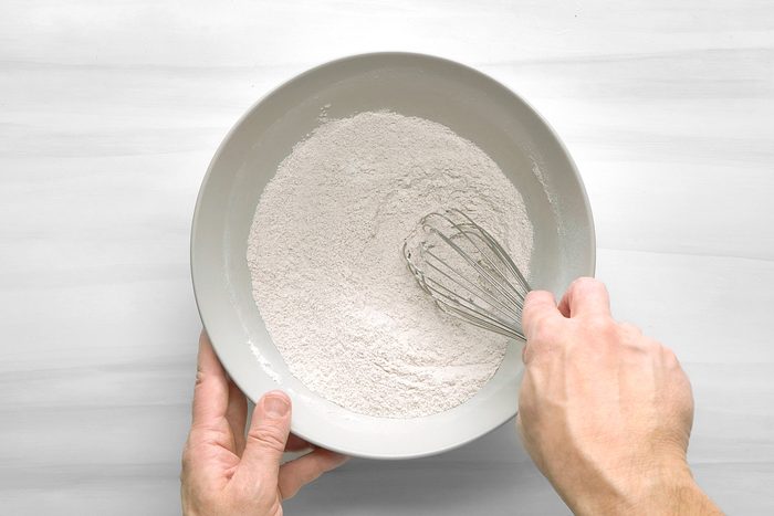 overhead shot of a person's hands holding a bowl with flour and a whisk; the bowl is gray and there is white flour in the bowl; the hands are holding the bowl and whisk; the background is a white surface