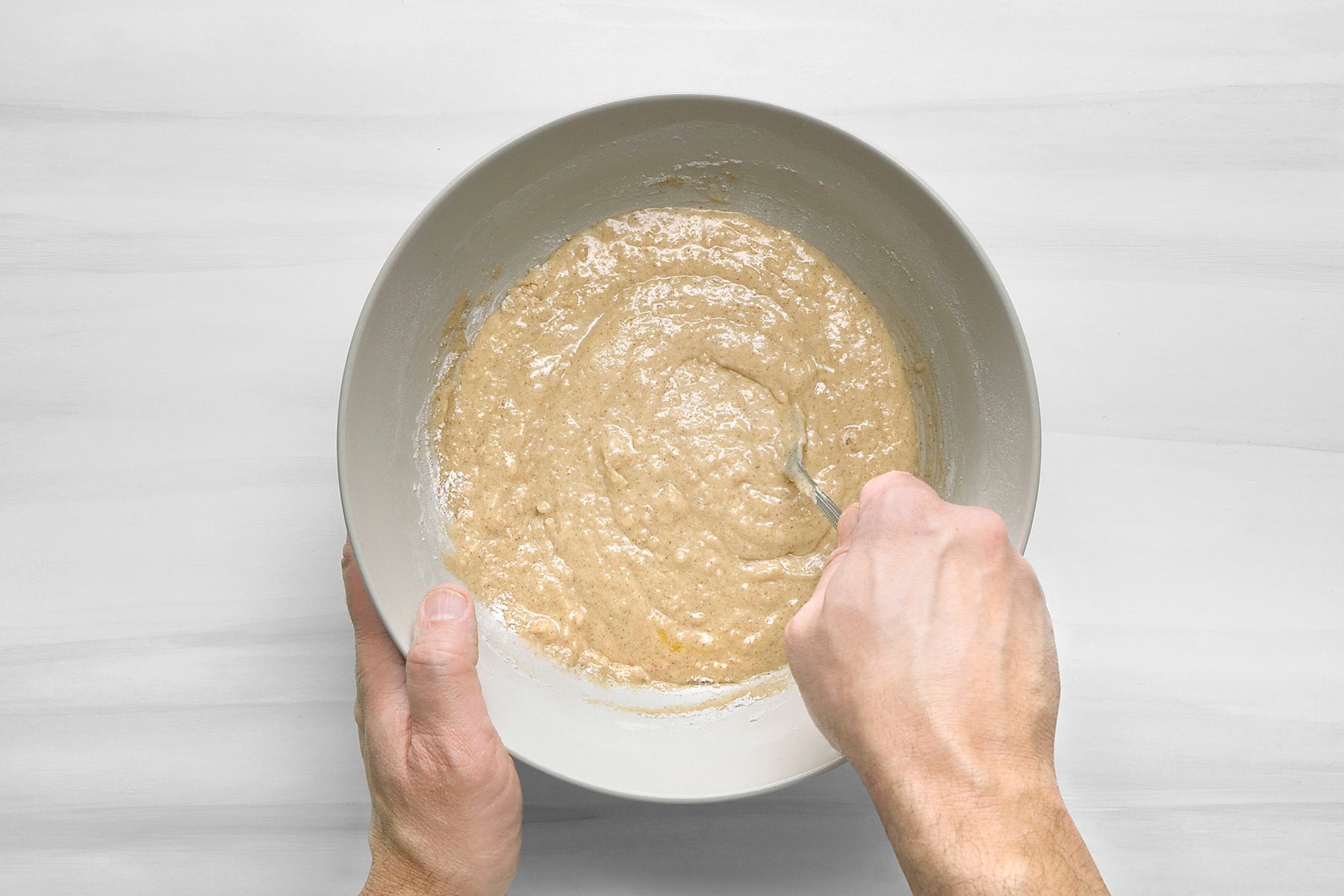 overhead shot of a bowl with a light brown batter inside, being mixed with a spoon, the bowl is on a white surface with faint wood grain pattern;