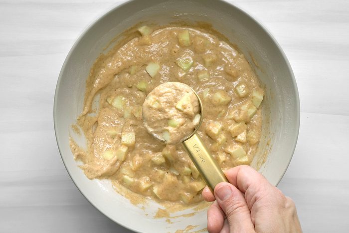 overhead shot of a bowl of batter with diced apples in it; a hand is holding a measuring cup that has some batter on it; the bowl and measuring cup are on a white surface