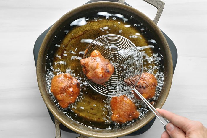 overhead shot of a hand holding a wire skimmer, with a metal handle, over a large pot of hot oil on the stove; the oil is bubbling rapidly, and there are four round golden-brown fried dough balls in the pot, the pot is sitting on a black mat and the background is a white countertop.