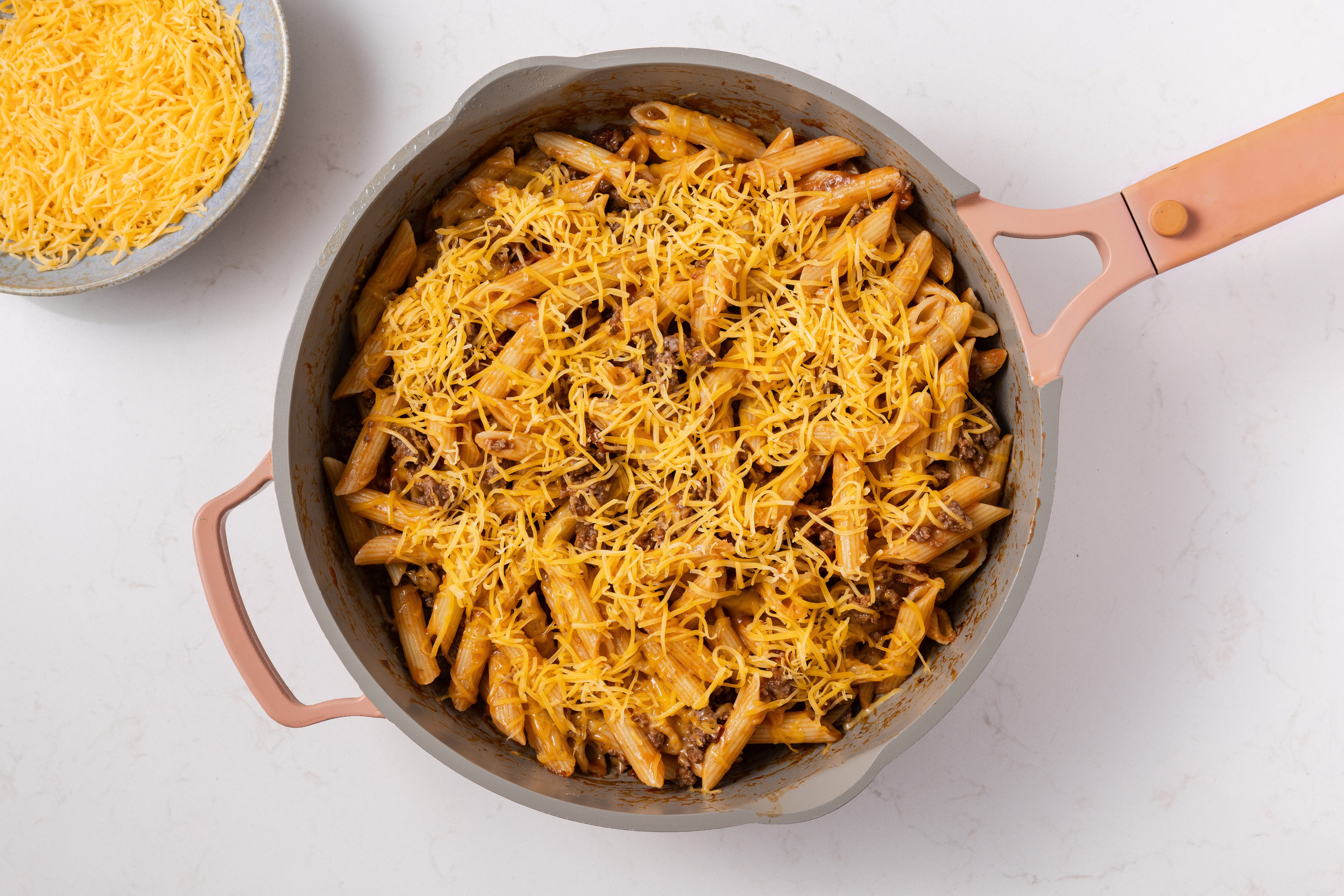 Bacon cheeseburger pasta being finalized with shredded cheddar cheese on top.