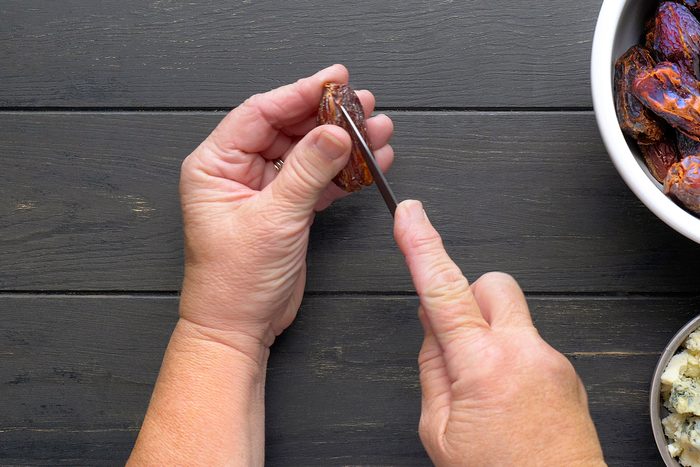 Close-up of hands using a knife to cut open a date on a dark wooden surface. A bowl of dates and another bowl with a cream-colored filling are visible nearby.