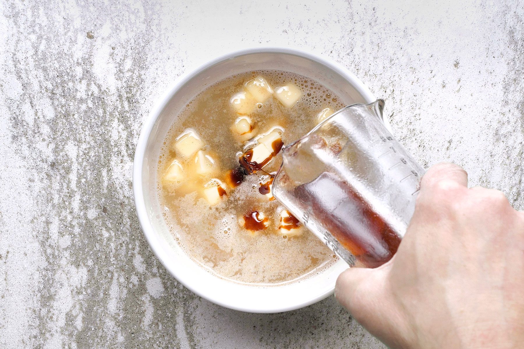 A hand pouring liquid from a glass measuring cup into a bowl containing a foamy mixture with small yellow cubed pieces, likely butter. The background is a textured light surface.