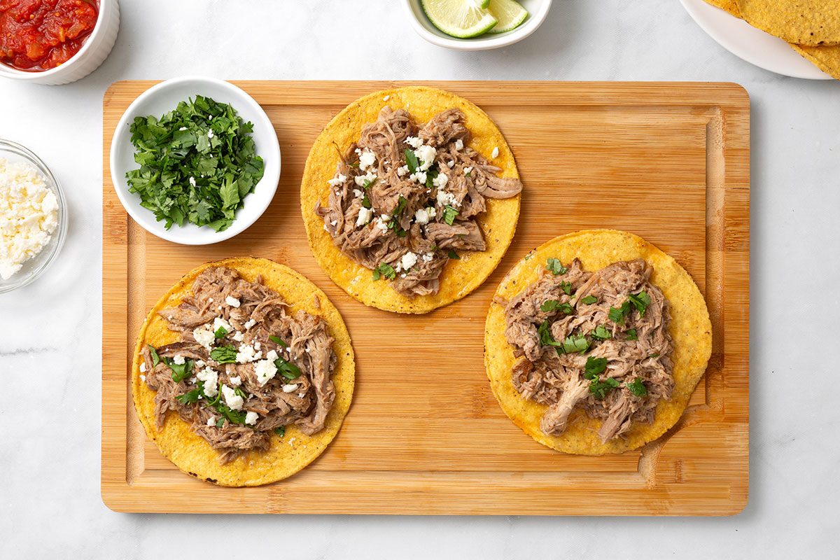 Tostada shells topped with shredded pork, chopped cilantro, crumbled queso fresco on a wooden cutting board for step four of Chipotle Carnitas recipe for Taste of Home